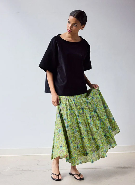 Woman wearing a relaxed black short-sleeve top paired with a flowing midi skirt in a green and blue paisley-inspired print. She is styled with black flip-flops and stands against a simple neutral backdrop while holding one side of the skirt.