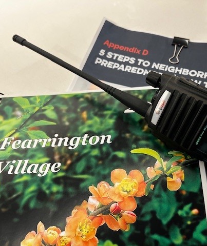 Handheld two-way radio resting on a Fearrington Village booklet with orange flowers on the cover, beside a printed neighborhood emergency preparedness guide.