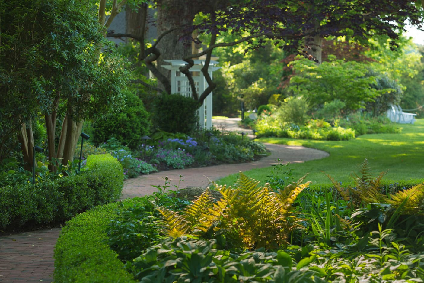 Lush landscaped gardens with a winding brick pathway, manicured shrubs, mature trees, and a white arbor in soft morning light. Fearrington Village.