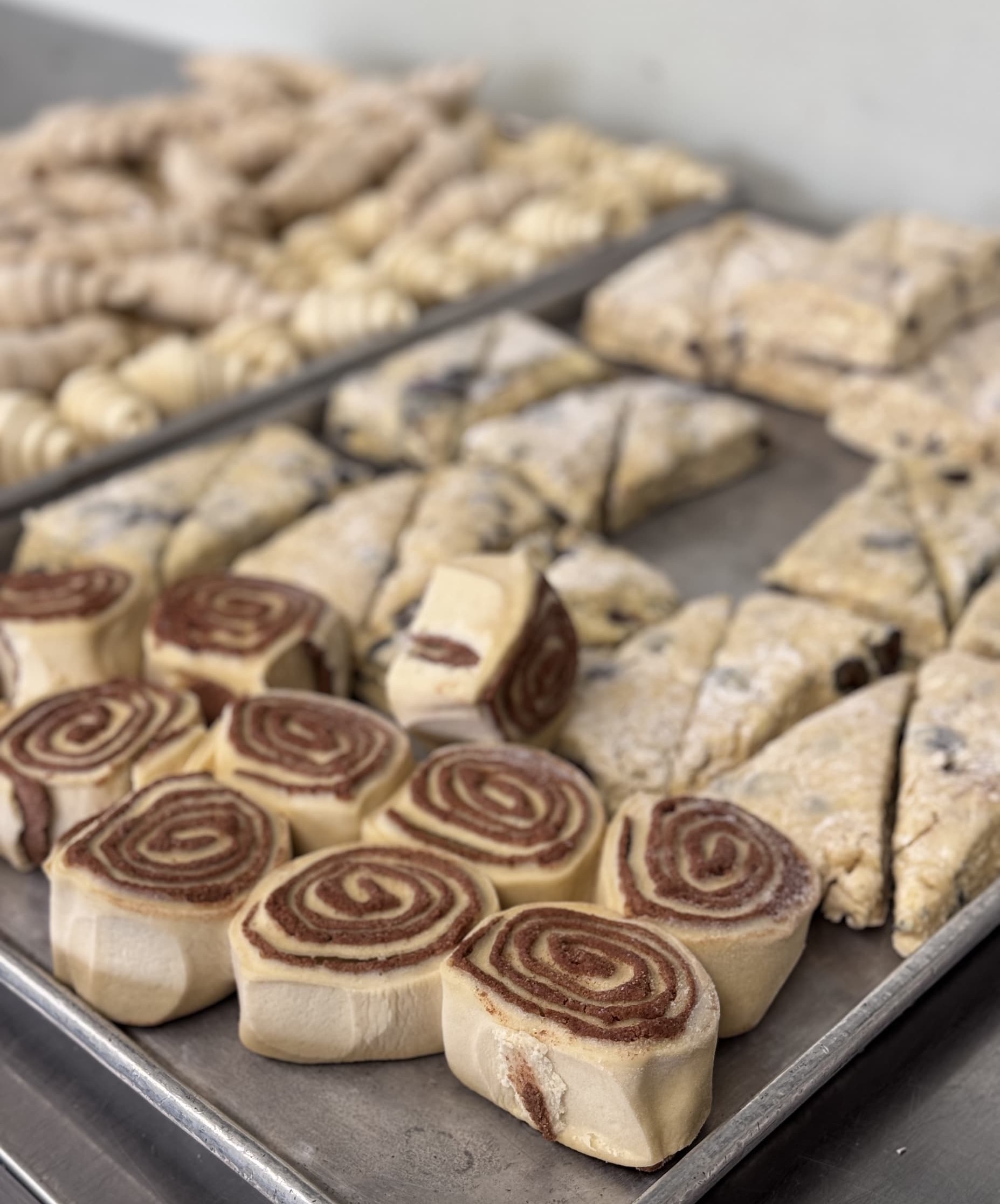 Unbaked pastries arranged on sheet pans, including cinnamon roll slices and triangular scones ready for the oven in a bakery kitchen.
