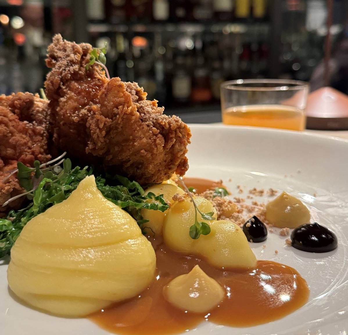 A plated dish featuring crispy fried chicken, piped mashed potatoes, sauces, and microgreens, with a bar full of bottles blurred in the background.
