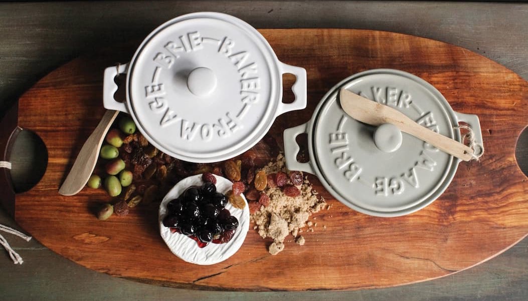 Top-down view of a wooden serving board with two small lidded ceramic dishes, a wheel of brie topped with dark fruit, green olives, and scattered dried fruit and crumbs, styled in a warm, rustic setting.