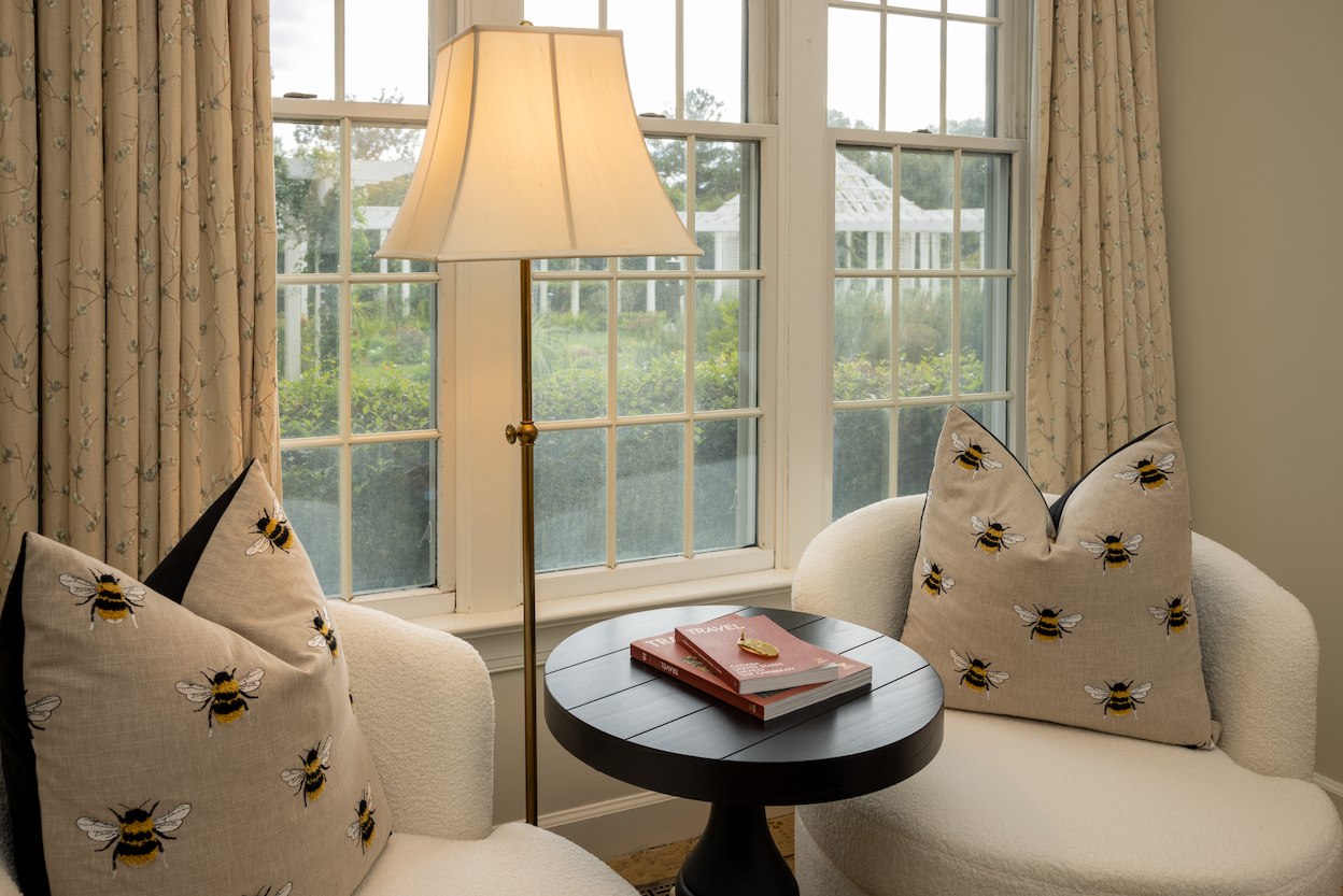 Cozy seating area at The Fearrington House Inn with cream chairs, embroidered bee pillows, a reading table and garden views through large windows. Fearrington Village.