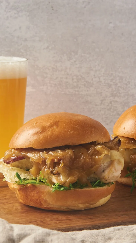Grilled chicken sandwich topped with caramelized onions and melted cheese on a brioche bun, served with fresh greens and paired with a pint of beer. Fearrington Village.