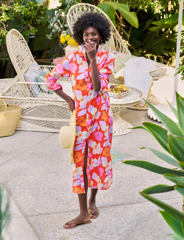 A smiling woman stands in a sunlit garden wearing a vibrant floral midi dress in red, pink, and orange tones. She holds a straw hat, wears flat sandals, and poses near white wicker lounge furniture surrounded by lush green plants.