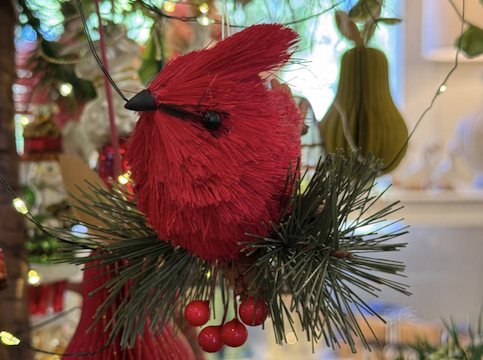 A red cardinal ornament made of textured material sits on artificial pine needles and red berries, surrounded by holiday lights and festive decorations. Fearrington Village