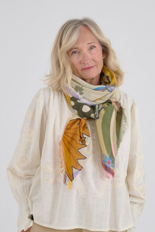 Woman wearing a light, embroidered blouse paired with a colorful patterned scarf in warm yellow, green, and soft pastel tones tied at the neck.