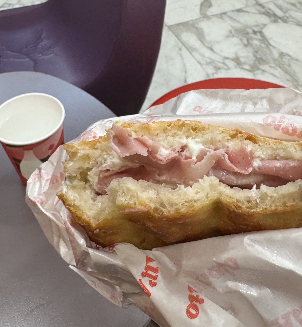 A close-up of a thick focaccia sandwich filled with thin slices of pink cured ham and creamy white cheese, partially wrapped in branded paper and resting on a café table beside a small paper cup.