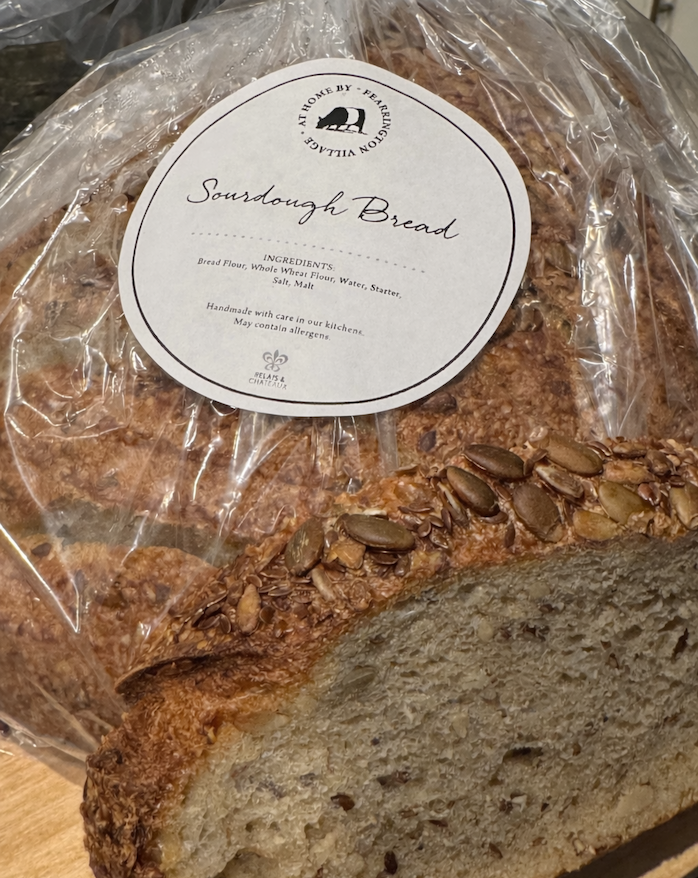 Loaf of seeded sourdough bread wrapped in plastic with a label reading “Sourdough Bread,” one slice cut to show the interior crumb.
