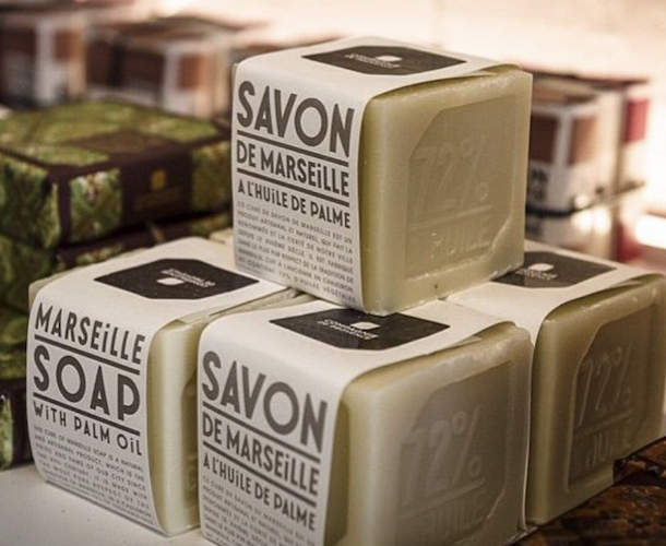 Close-up of stacked Marseille soap cubes wrapped in clean white labels, highlighting palm oil content, set against a softly lit display of artisanal soaps in the background.