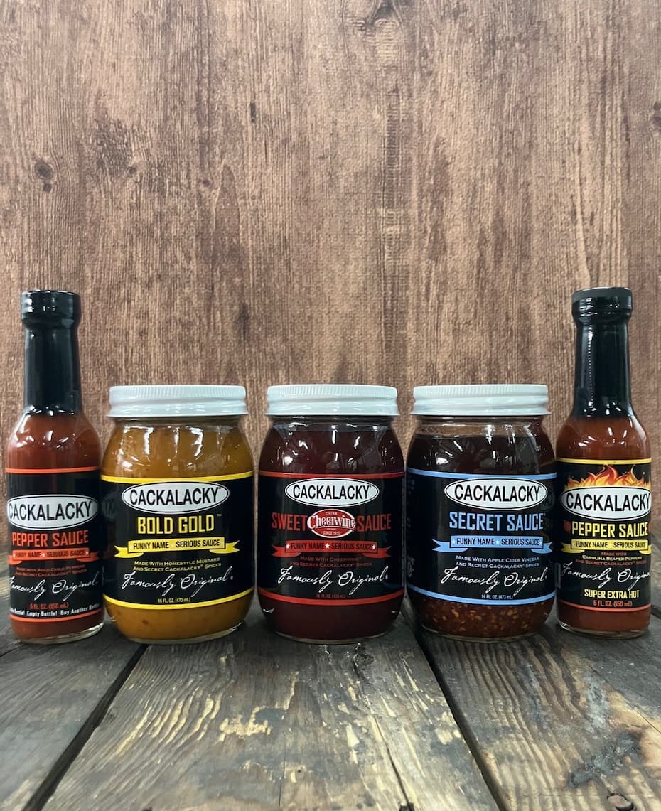 A collection of Cackalacky sauces and condiments is displayed on a rustic wooden table against a weathered wood backdrop. The lineup includes Original Pepper Sauce, Bold Gold mustard, Sweet Cheerwine Sauce, Secret Sauce, and Super Extra Hot Pepper Sauce. The jars and bottles feature bold black labels with colorful accents, showcasing the North Carolina brand’s playful “Funny Name. Serious Sauce.” tagline. The arrangement highlights a variety of flavors ranging from sweet and tangy to intensely spicy.