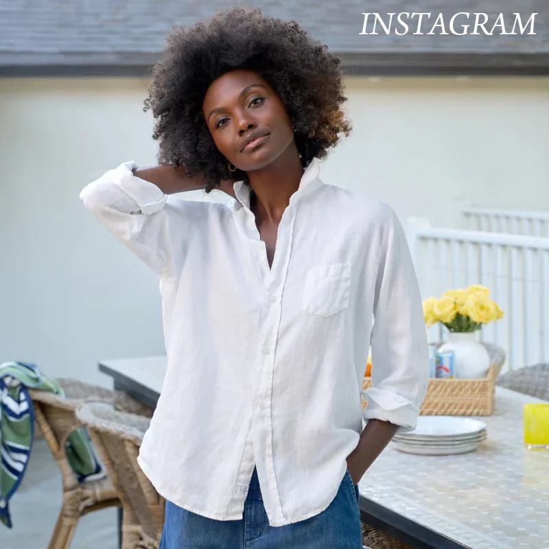 Woman standing outdoors near a patio table, wearing a relaxed white button-down shirt with sleeves rolled and dark jeans. She poses casually with one hand in her pocket and the other behind her neck, with wicker chairs and flowers in the background.