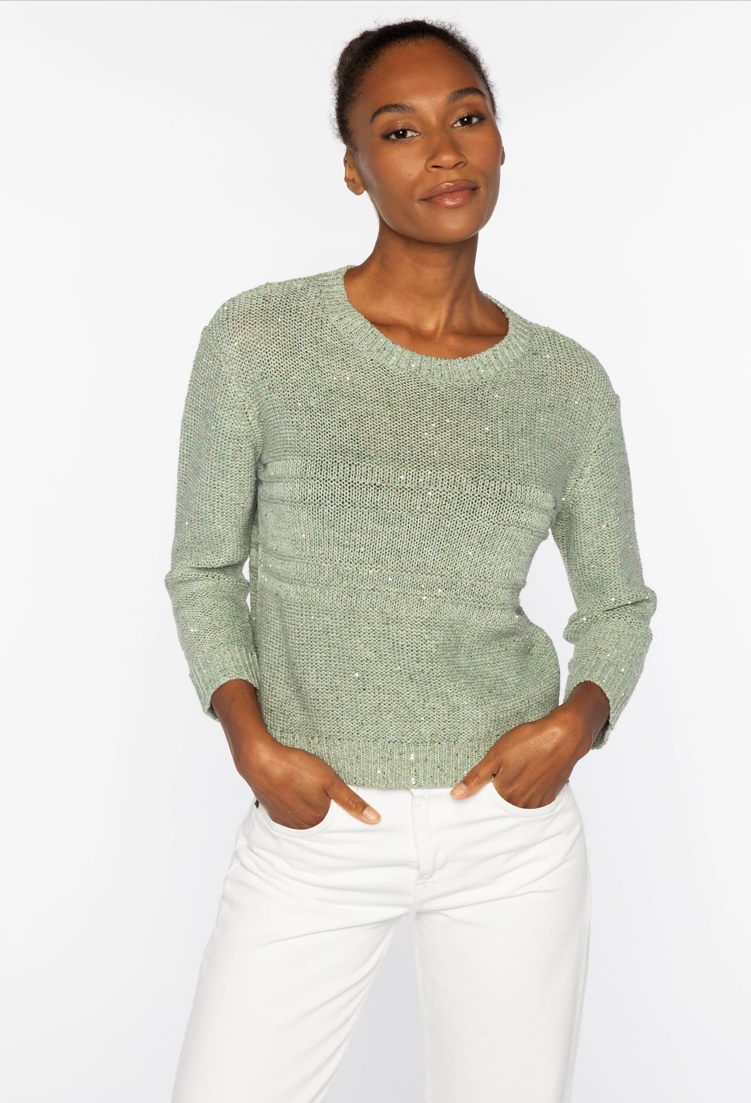 Woman wearing a soft sage-green knit sweater and light trousers, standing against a neutral background with hands in her pockets.