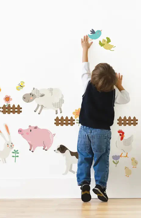 A young child reaches up toward colorful removable farm animal wall decals, including sheep, pigs, chickens, birds, and a fence, arranged playfully on a white wall.
