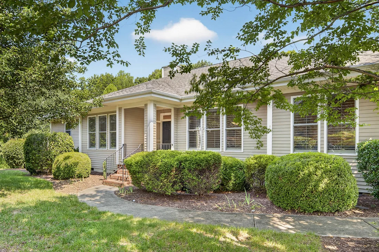Single-story cottage with large windows, manicured shrubs, mature trees, and a curved front walkway. Fearrington Village.