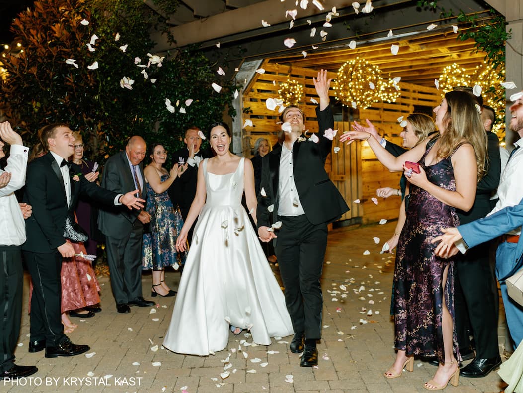 Bride and groom walking hand in hand outside The Fearrington Barn through guests tossing white flower petals, outdoors at night with warm string lights overhead.