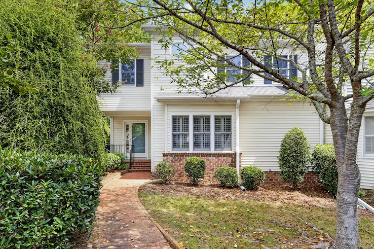 Two-story white home with black shutters, a brick walkway, mature trees, and neatly landscaped shrubs. Fearrington Village.