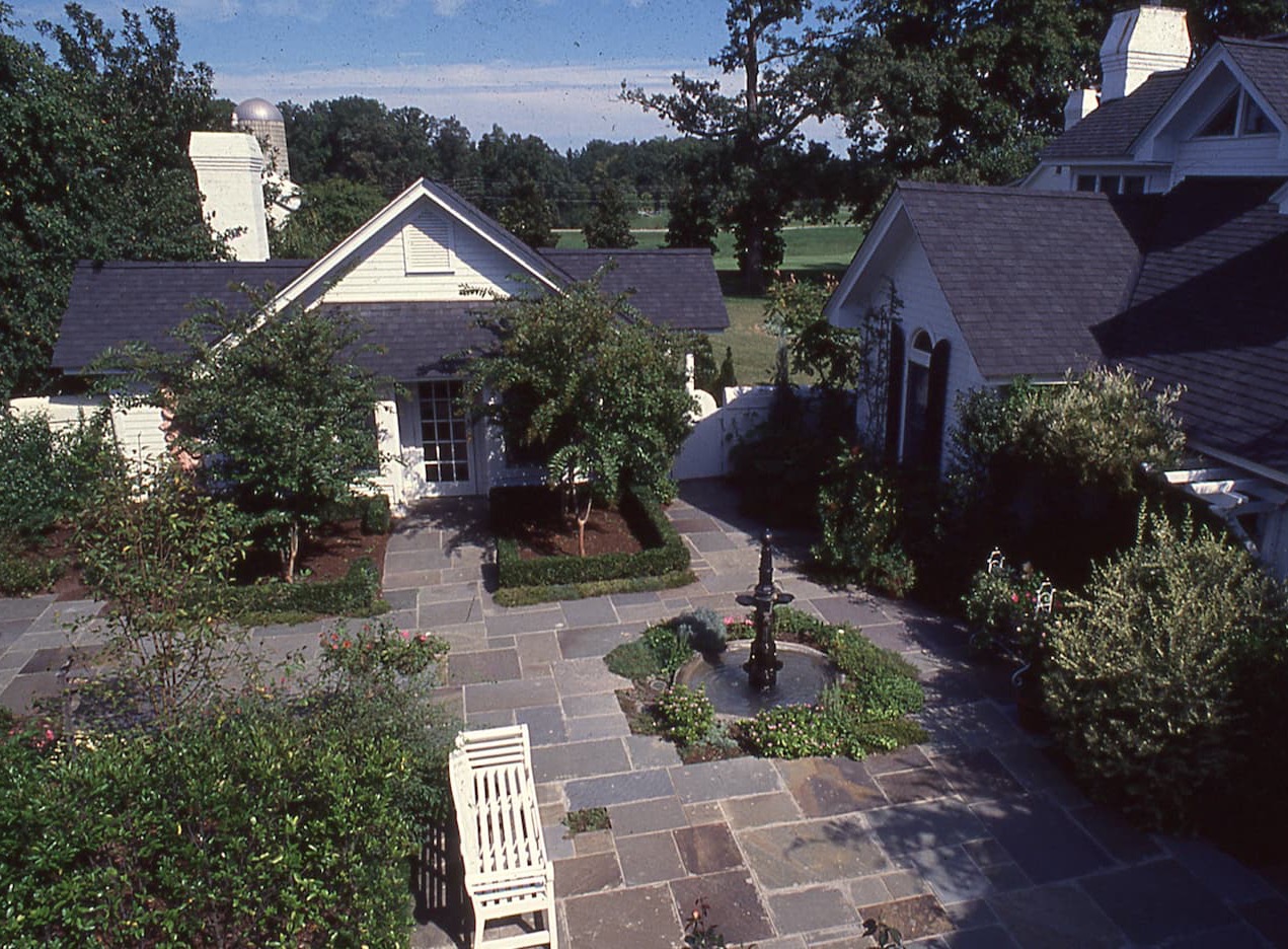 Historic photo of the courtyard at the Fearrington House Inn, showing a brick-and-stone terrace with a central fountain, garden beds, and white cottage buildings surrounded by mature trees and lawns.