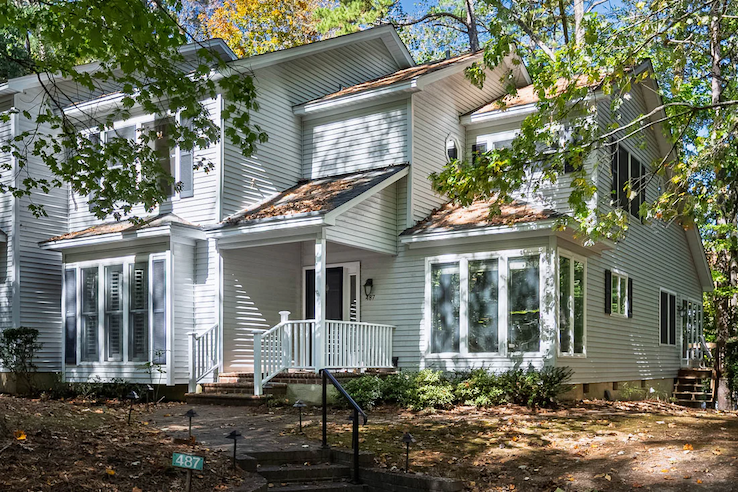 A light-colored two-story townhouse with a covered front porch and large windows sits among mature trees, with dappled sunlight across the yard.