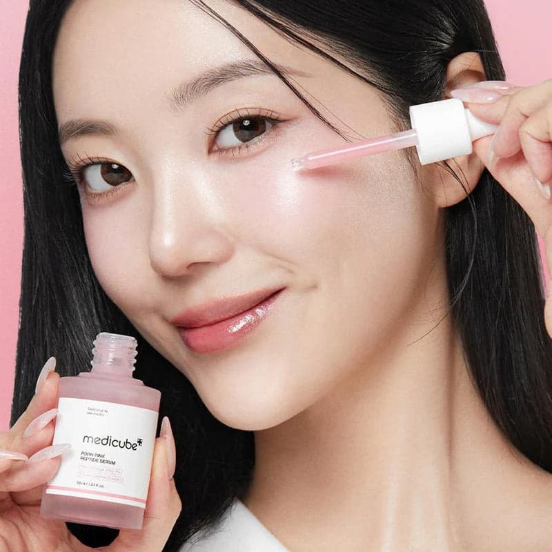 Woman applying Medicube PDRN Pink Peptide Serum with a dropper against a pink background. Fearrington Village.