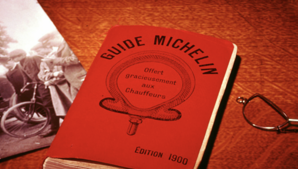 Vintage 1900 Michelin Guide with its distinctive red cover, displayed alongside an early motoring photograph and eyeglasses. Fearrington Village.