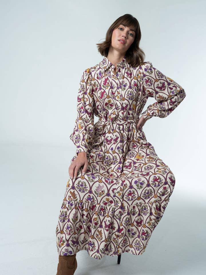 A woman models a long belted shirt dress in an intricate botanical print of plum, purple, gold and green on a cream background. Fearrington Village.