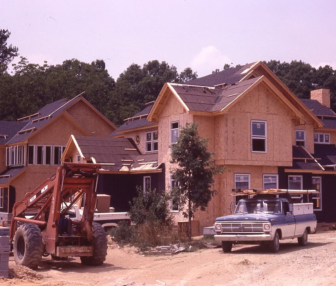 Early construction of the Fearrington Inn, showing wood-framed buildings with unfinished siding and roofing, a front loader, and a pickup truck on a dirt worksite surrounded by trees.