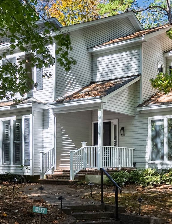 Exterior of a light gray two-story townhome with white trim, a small covered porch and railing, brick steps, and mature trees casting dappled shade across the front yard.