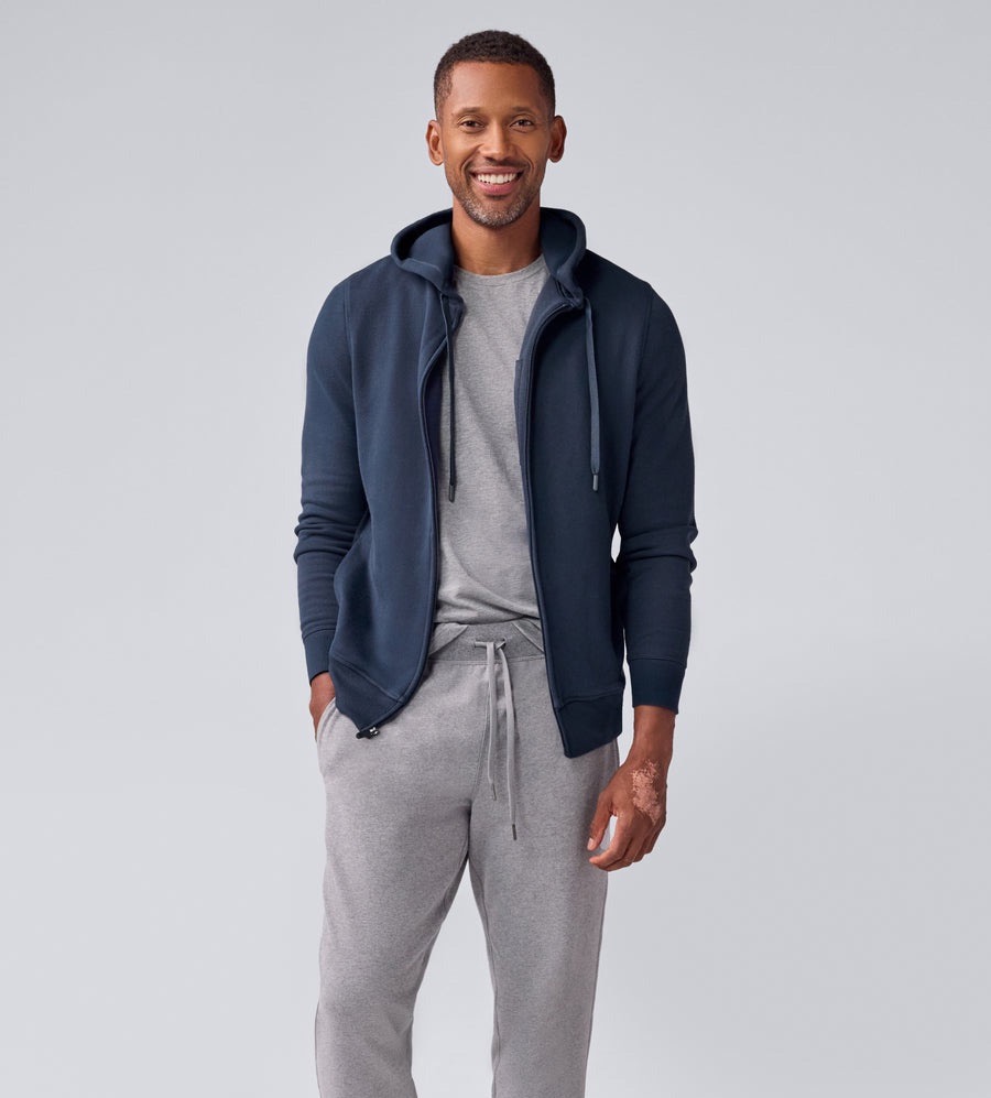 Man wearing a navy zip hoodie over a gray t-shirt with gray jogger pants stands smiling against a light gray background.
