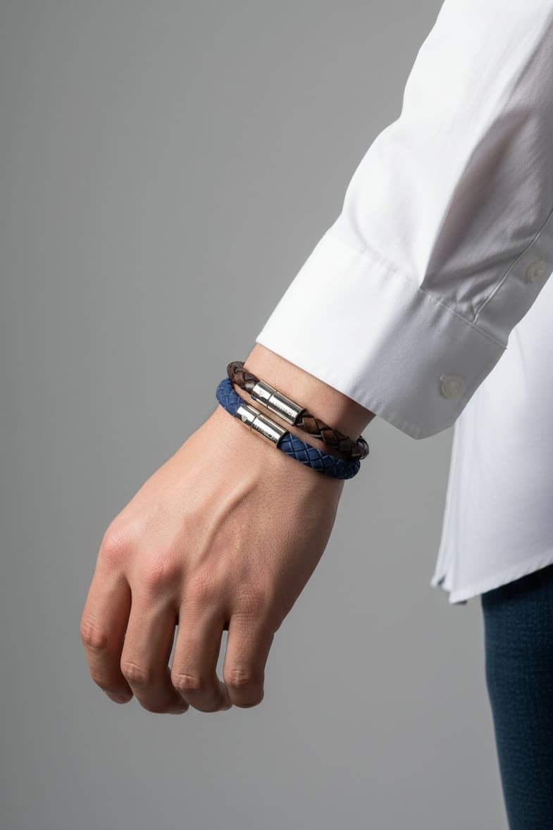 Close-up of a person’s wrist wearing two braided leather bracelets—one navy and one brown—with polished metal clasps, styled with a white button-down shirt against a neutral background.