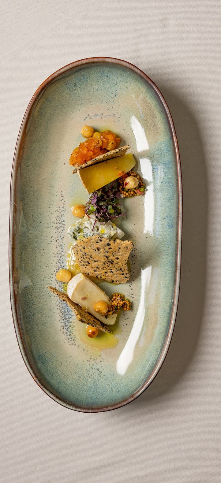 Artfully plated cheese course featuring layered cheeses, seeded crisps, mustard seeds, microgreens, and small dollops of purée, arranged on an elongated ceramic platter with a refined, modern presentation.