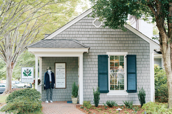 A small boutique with gray shingles, blue shutters, and a mannequin dressed in casual clothes near the entrance. Green plants and trees surround the shop, and a sign reads "Thistle & Oak. Fearrington Village