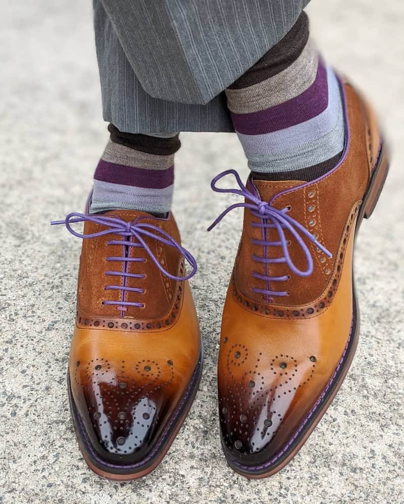 A pair of tan and brown polished brogue shoes with purple laces worn with striped purple and gray socks on a concrete surface.