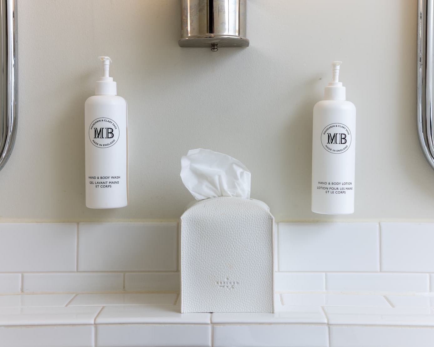 Minimal bathroom setup with two white pump bottles for hand wash and body lotion mounted on a wall above white tile, centered around a textured tissue box beneath a chrome fixture.