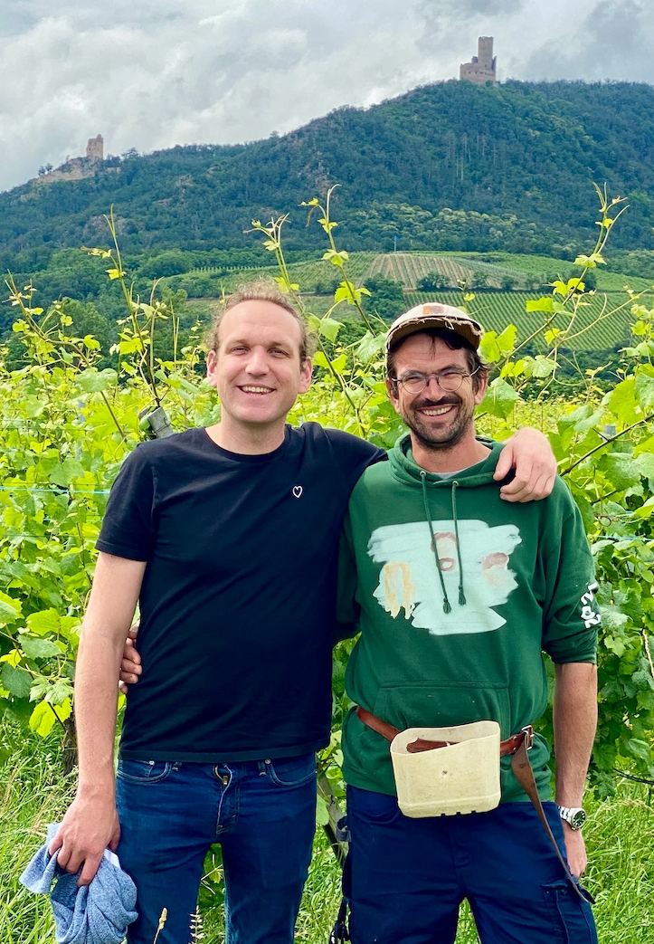 Two men standing arm-in-arm in a lush vineyard, smiling at the camera, with rolling green hills and a stone tower on a mountaintop in the background under a cloudy sky.