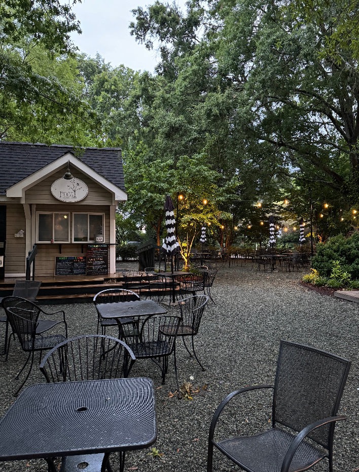 A quaint outdoor café with a small wooden building featuring a menu board and sign that reads "Food". The seating area has wrought-iron tables and chairs on a gravel ground. Black-and-white striped umbrellas are scattered around. Tall green trees surround the serene setting. Fearrington Village