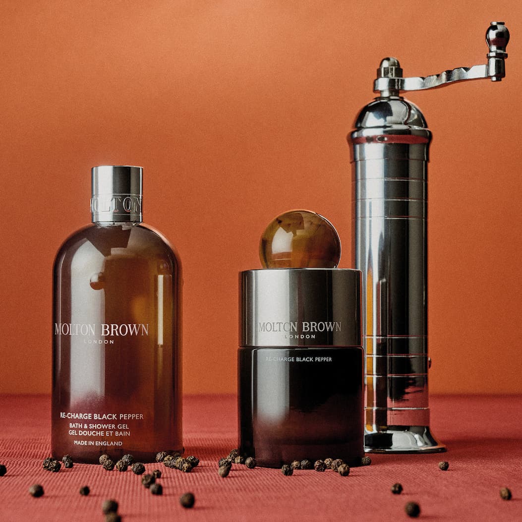 Molton Brown Re-charge Black Pepper bath and shower gel and fragrance displayed beside a pepper grinder on a warm orange background.