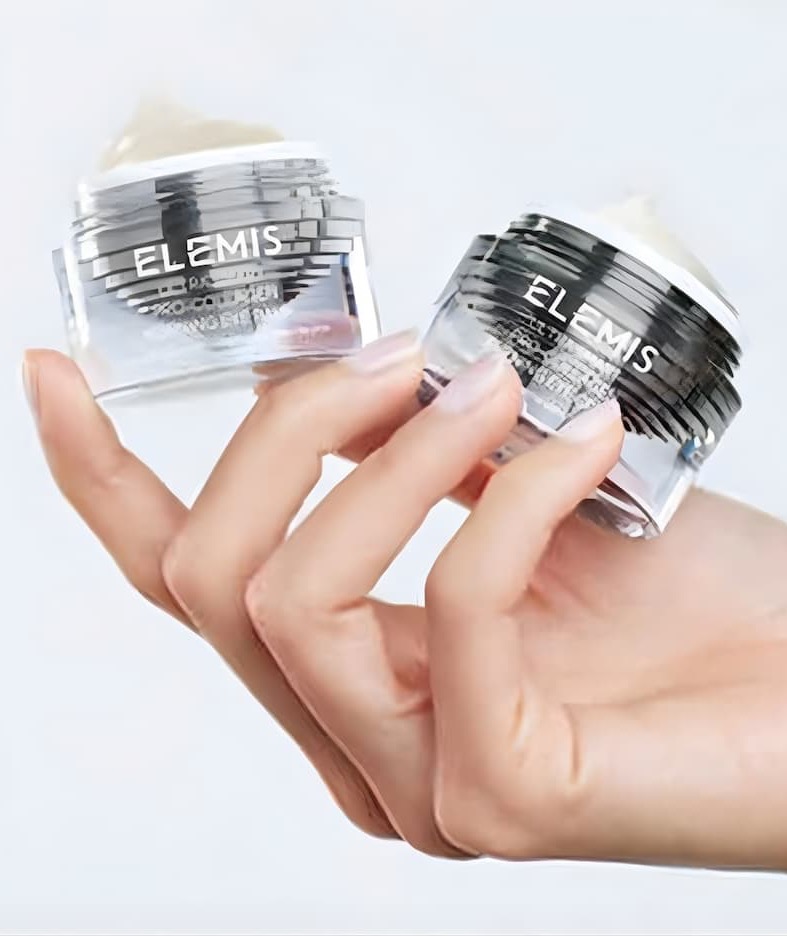 Hand holding two clear jars of Elemis face cream with black lids against a white background.