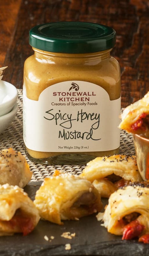 Jar of Stonewall Kitchen Spicy Honey Mustard on a wooden surface, surrounded by bite-sized pastry appetizers and a small dipping bowl.