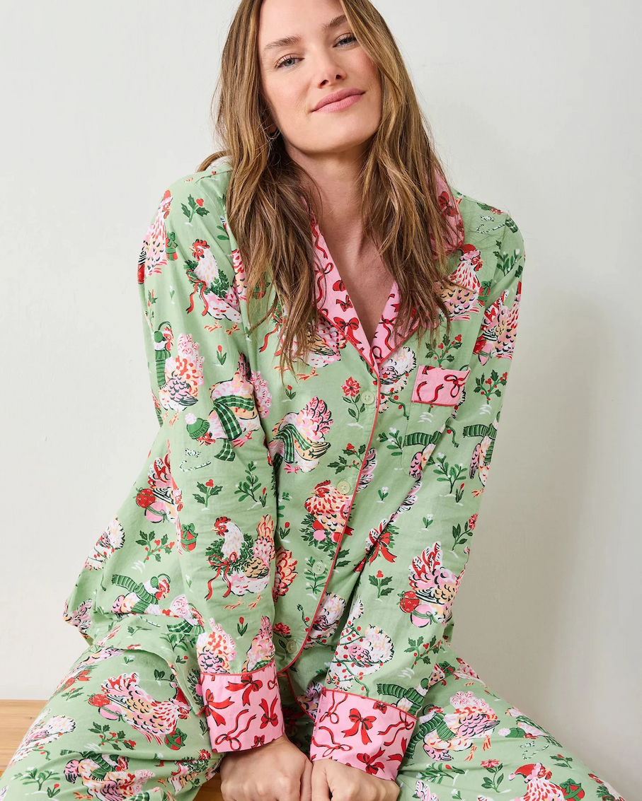 Woman with long light brown hair wearing a green pajama set printed with whimsical birds and florals, featuring pink contrast trim and cuffs, seated casually against a light wall.