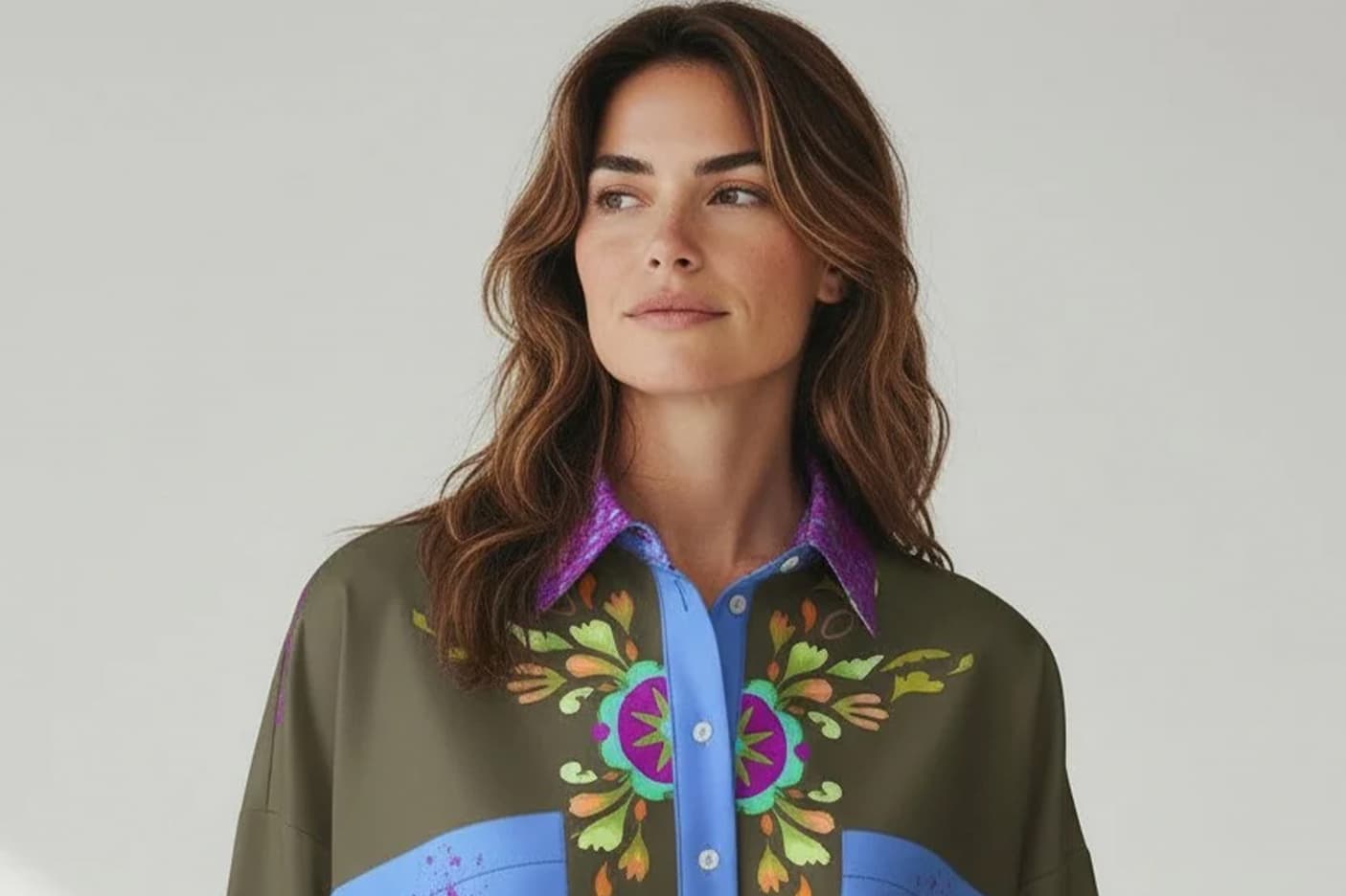 A woman models a statement blouse with a bold, symmetrical floral motif in vibrant greens, purples, and blues, accented by a contrasting blue placket and shimmering purple collar for a polished, artistic look.