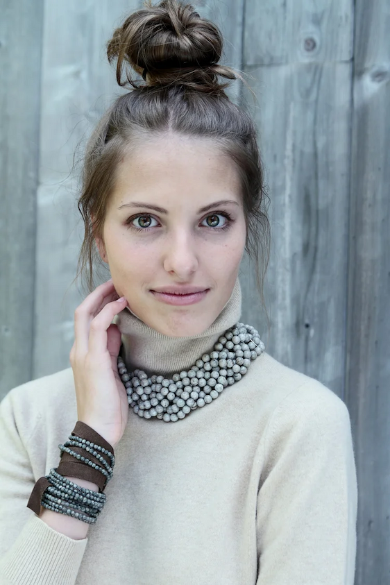 Portrait of a woman with light skin and green eyes, wearing a beige turtleneck sweater and layered gray beaded necklace and bracelets. Her hair is styled in a high messy bun, and she gently touches her neck while looking at the camera, set against a weathered wooden backdrop.