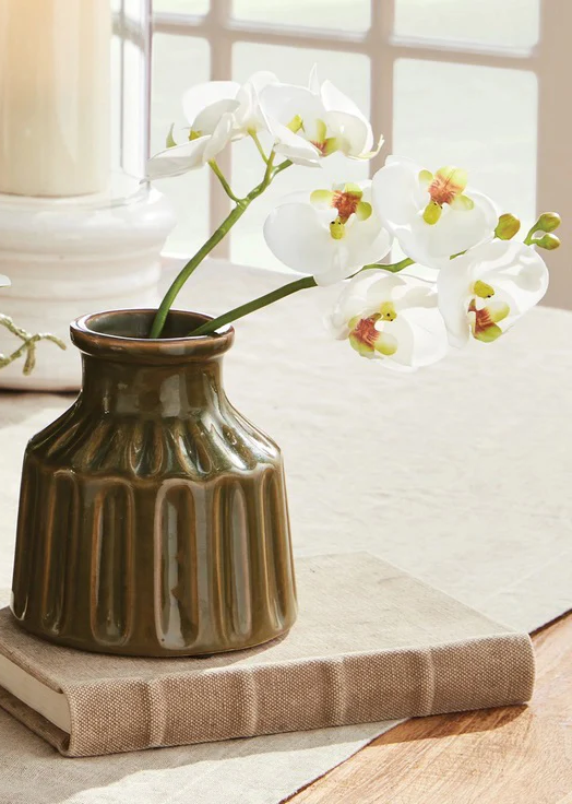 White orchid stems arranged in a textured olive ceramic vase resting on a linen-covered book beside a sunlit window. Soft natural light and warm neutral tones create a calm, elegant tabletop vignette.