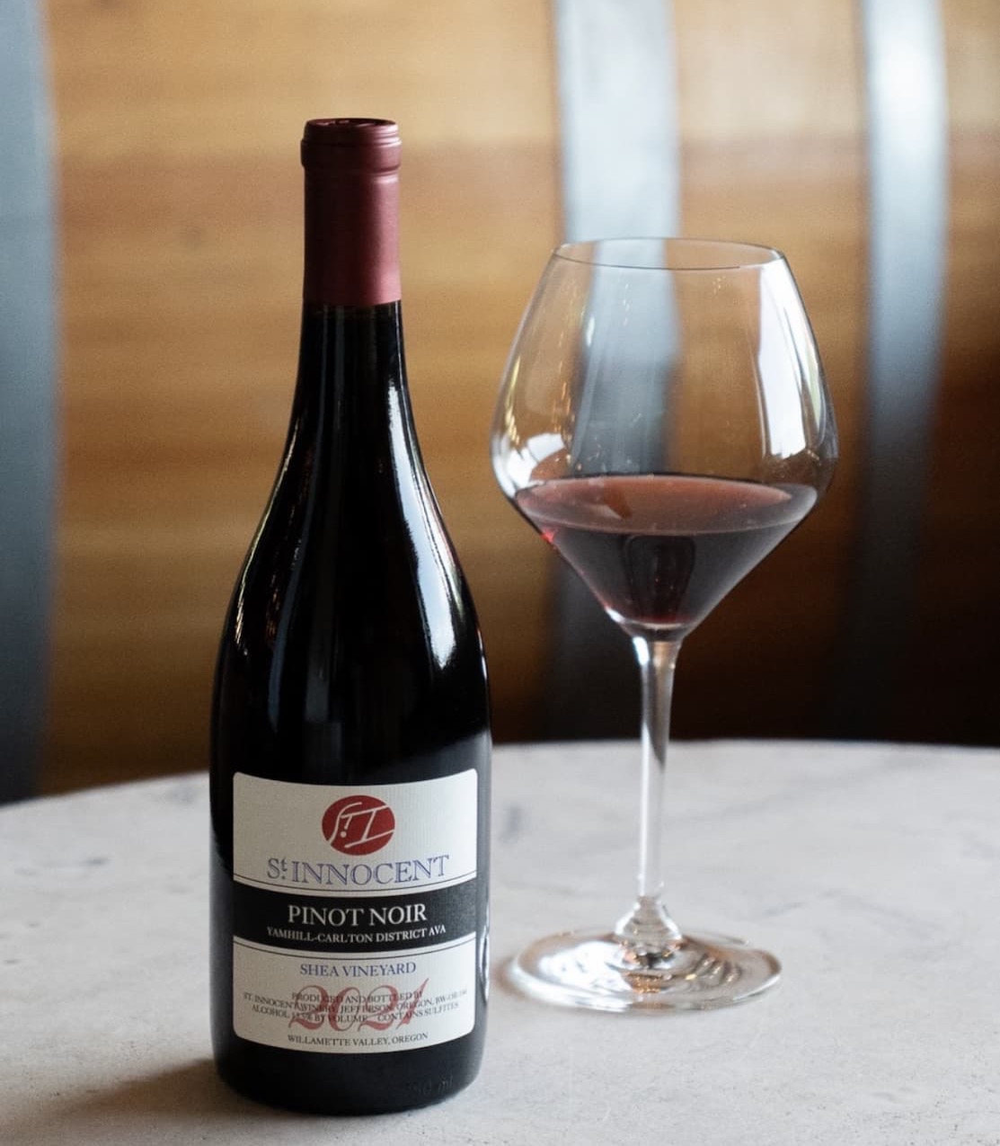 A bottle of St. Innocent Pinot Noir from Oregon beside a partially filled glass of red wine on a light tabletop, with a softly blurred warm-toned background.