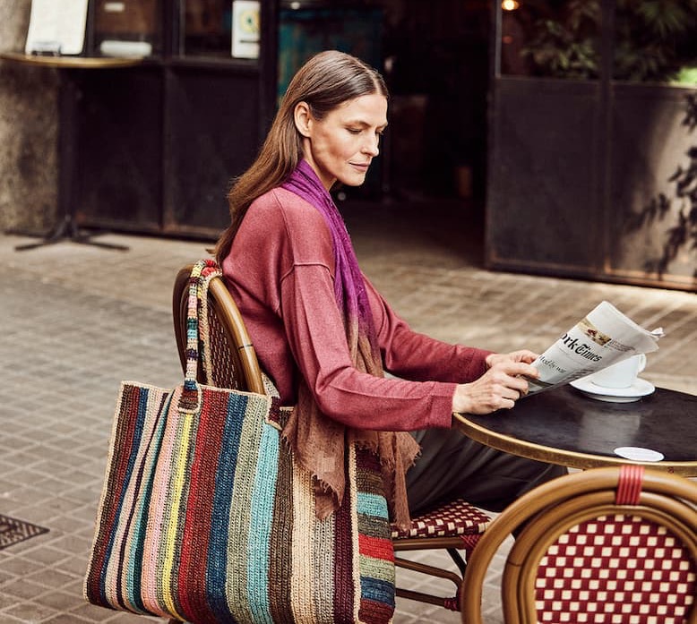 A woman sits at an outdoor café table reading a newspaper while enjoying a quiet moment. Beside her, a large woven tote bag with multicolored vertical stripes rests against a bistro chair. The textured natural-fiber bag adds a vibrant touch to her relaxed outfit, making it an ideal companion for market visits, travel, or everyday errands.