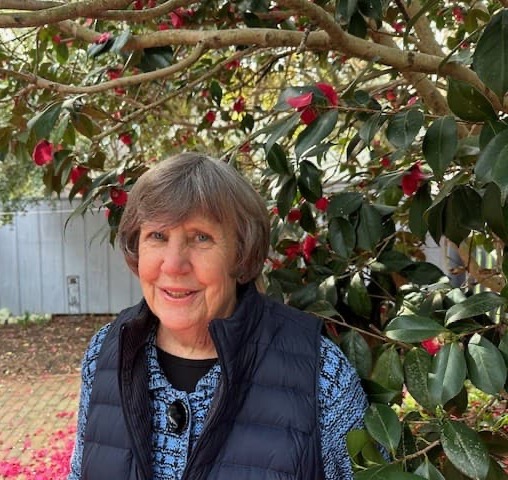 An older woman stands outdoors beneath a flowering tree, wearing a dark vest over a patterned top and smiling warmly.