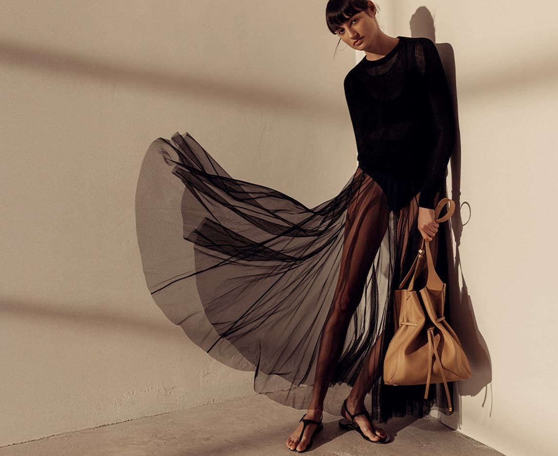Woman wearing a flowing black sheer skirt and knit top, carrying a tan leather handbag in warm sunlight. Fearrington Village.
