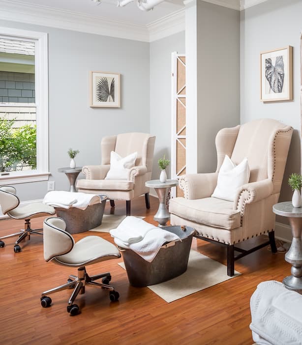 Bright, serene spa pedicure room with two upholstered armchairs, foot basins with neatly folded white towels, rolling stools, and soft neutral décor accented by framed botanical prints and warm wood floors.