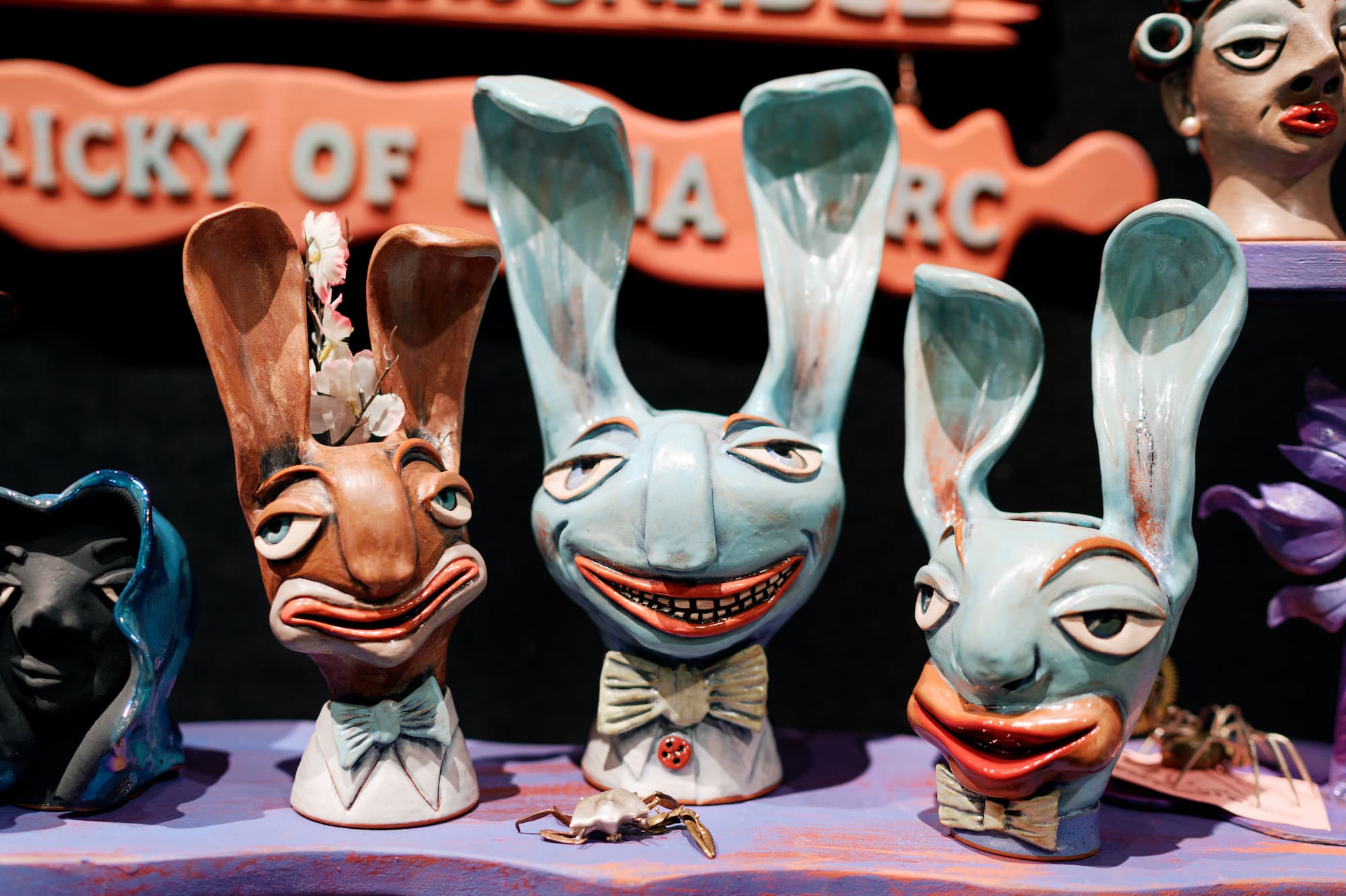 A display of whimsical ceramic rabbit sculptures with exaggerated facial expressions sits on a table at an art market. The hand-painted rabbits feature tall ears, bow ties, and playful details in shades of blue, brown, and cream, with a vintage-style sign blurred in the background.