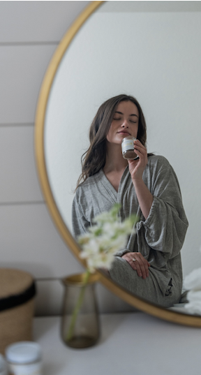 Woman in a soft gray lounge set sits beside a round mirror, holding a candle near her face with eyes closed. The serene bedroom scene features muted tones, natural light, and blurred flowers in the foreground, creating a calm, spa-like atmosphere.